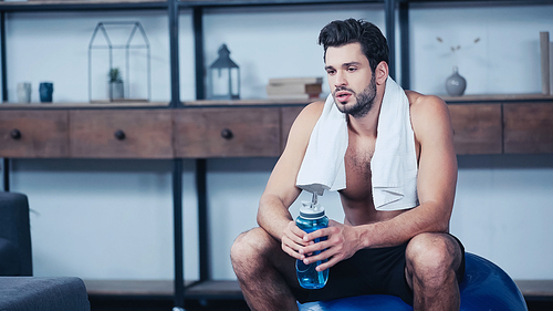 tired young sportsman with towel sitting on fitness ball and holding sports bottle