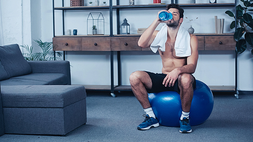 full length of tired sportsman with towel sitting on fitness ball and holding sports bottle while drinking water
