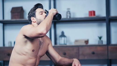 muscular sportsman holding sports bottle and drinking water at home