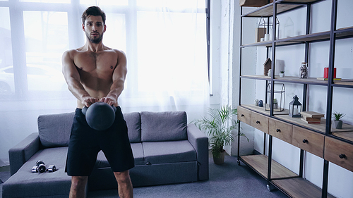 young muscular sportsman in shorts training with heavy kettlebell near sofa