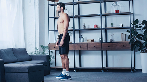 full length of muscular man in shorts standing near sofa