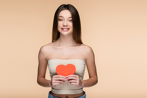 happy woman in crop top with bare shoulders holding red paper heart isolated on beige