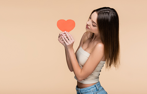 young woman in crop top with bare shoulders looking at red paper heart isolated on beige