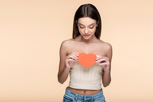 brunette woman in crop top with bare shoulders looking at red paper heart isolated on beige