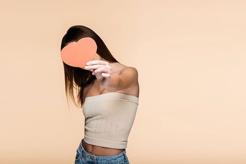 brunette young woman in crop top with bare shoulders covering face with red paper heart isolated on beige