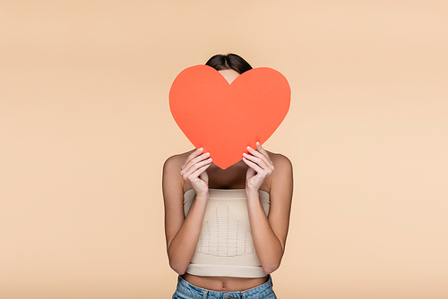 young woman in crop top with bare shoulders covering face with red paper heart isolated on beige