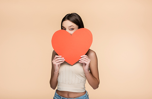 young woman in crop top covering face with red paper heart isolated on beige