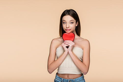 brunette young woman in crop top holding red heart-shaped metallic box isolated on beige