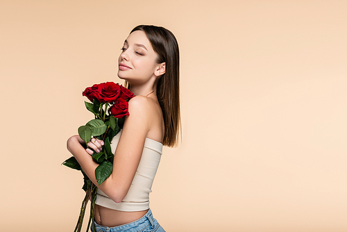happy young woman with closed eyes holding red roses isolated on beige