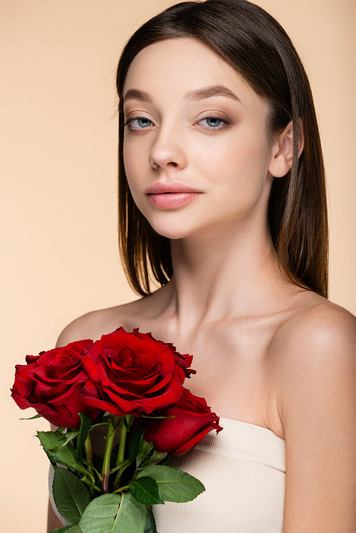 brunette young woman with bare shoulders holding red roses isolated on beige