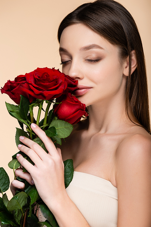 brunette young woman with closed eyes smelling red roses isolated on beige