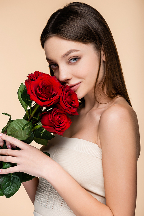 pleased young woman with bare shoulders smelling red roses isolated on beige