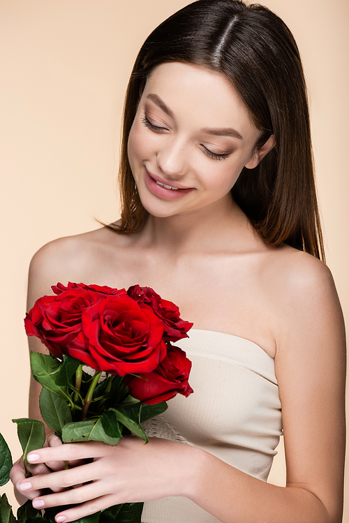 happy woman with bare shoulders looking at bouquet of red roses isolated on beige