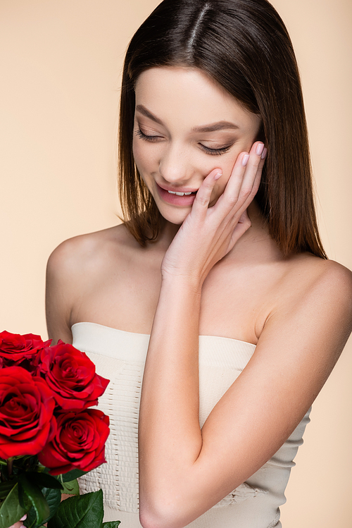 cheerful woman with bare shoulders looking at bouquet of red roses isolated on beige