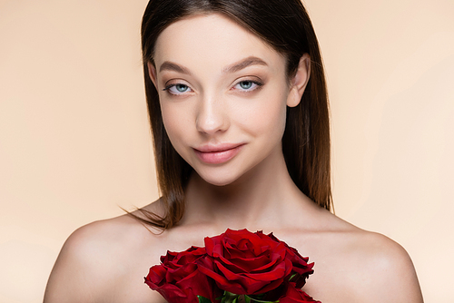 young woman with bare shoulders near bouquet of red roses isolated on beige