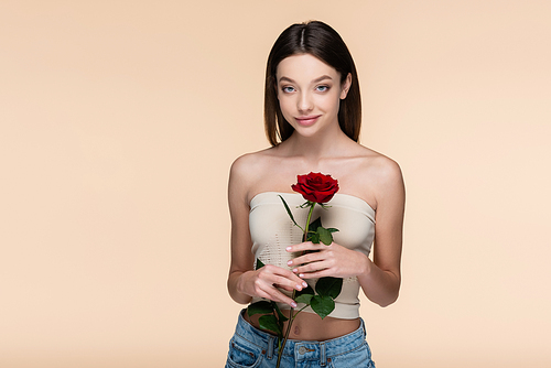 pleased young woman with bare shoulders holding red rose isolated on beige