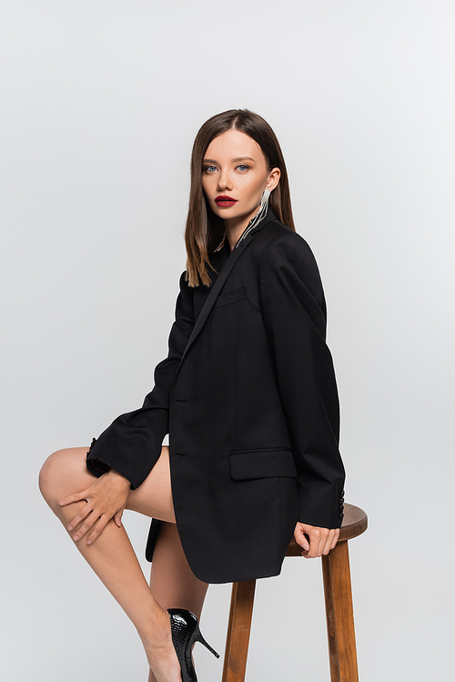 seductive brunette woman with naked legs, wearing black blazer,  on high stool isolated on grey