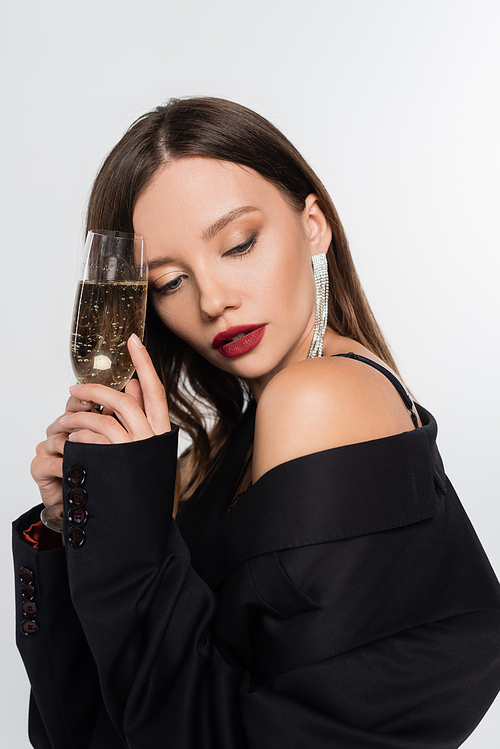 seductive brunette woman in black blazer holding glass of champagne isolated on grey