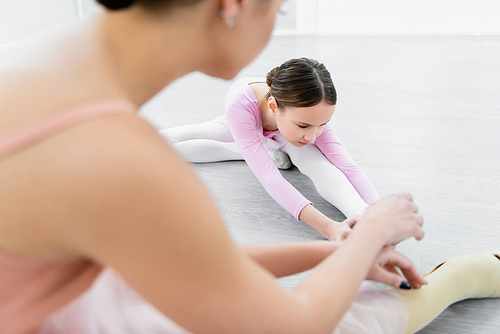 girl stretching on floor in ballet school near blurred dance teacher