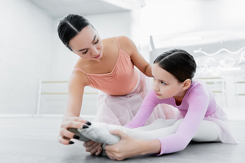 preteen girl looking at dance teacher while stretching on floor in ballet school