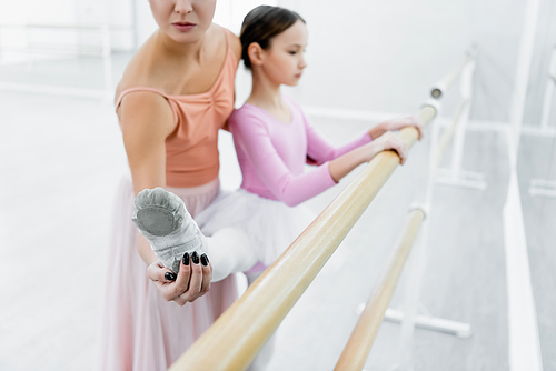 ballet teacher helping girl stretching at barre in dance studio, blurred background