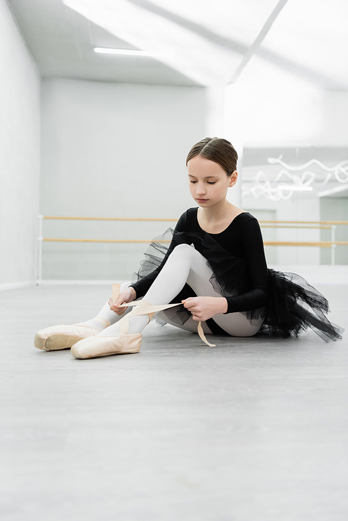 child in black ballet costume tying pointe shoe on floor in dancing hall