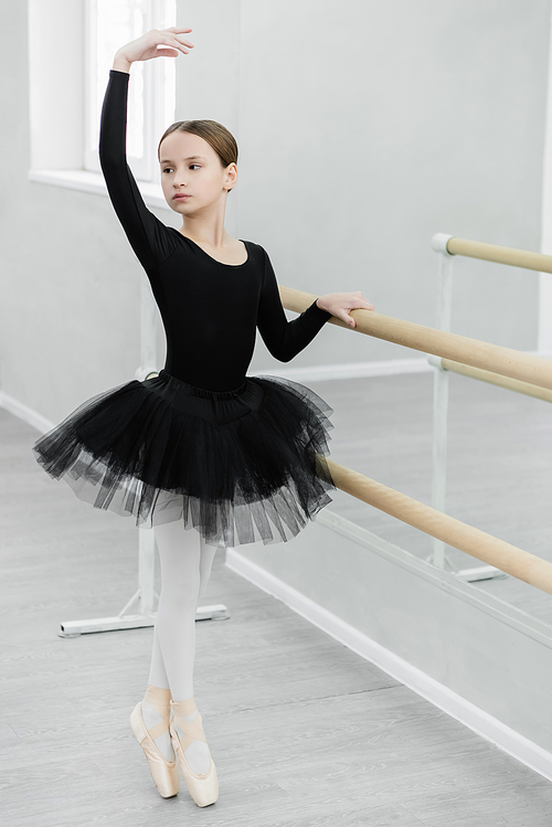 slim girl in black tutu standing on toe while training at barre in dancing studio