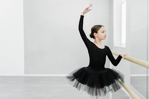 preteen girl in black tutu looking in mirror while training at barre in ballet studio