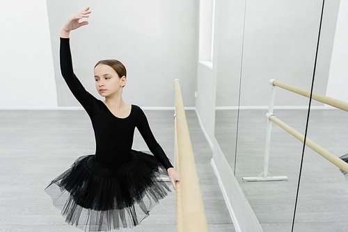 graceful girl in black tutu training with raised hand at barre near mirrors