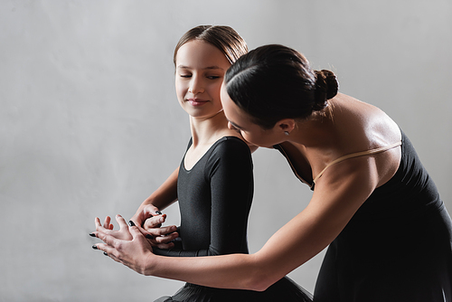 young ballet teacher touching hands of girl during dance lesson on grey background