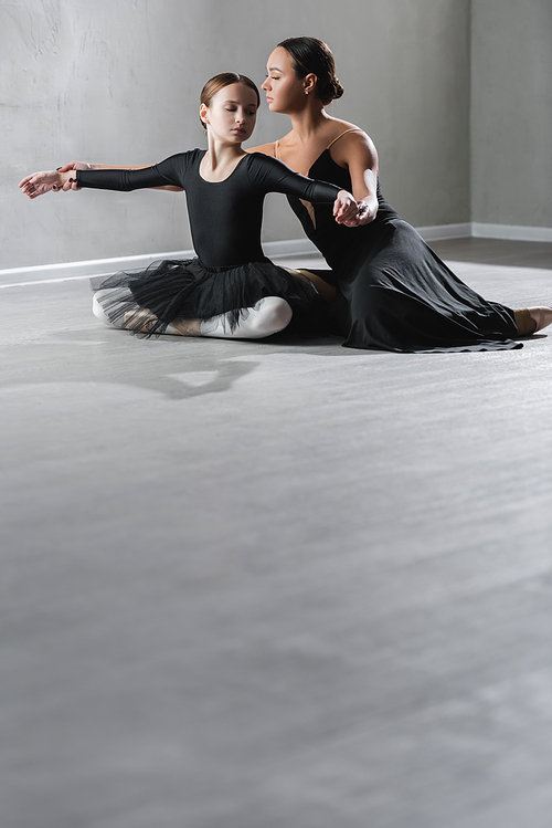 girl in black tutu sitting with crossed legs near ballet teacher in studio