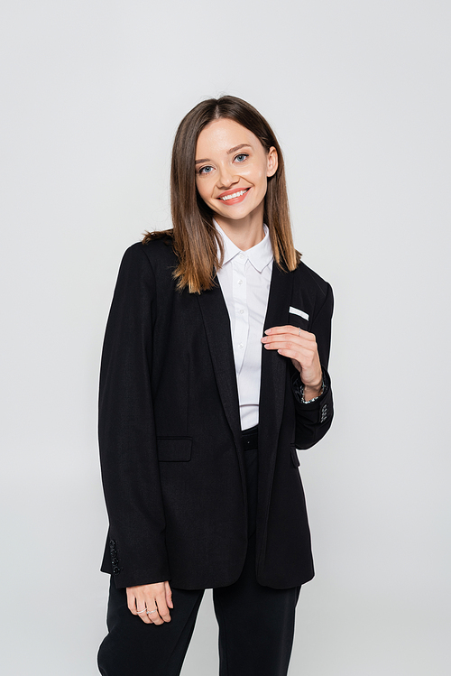 happy and stylish woman in suit smiling isolated on grey