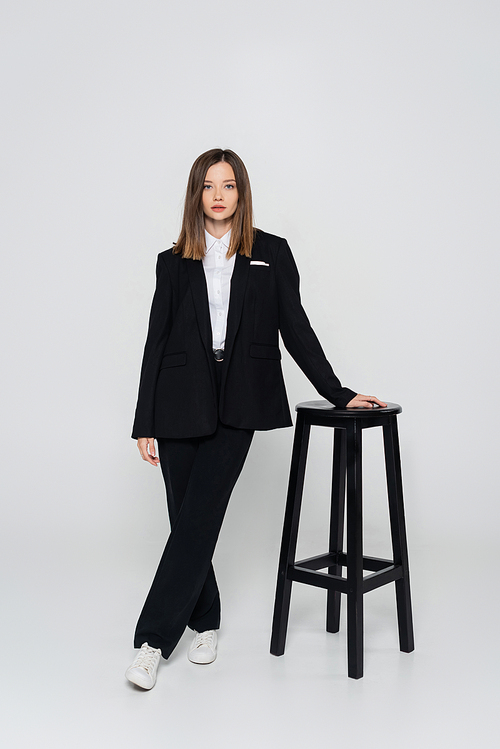full length of young stylish woman in suit posing near chair on grey
