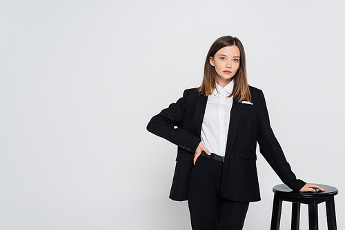 young stylish woman in suit posing with hand on hip near chair isolated on grey