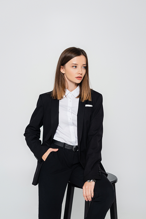 young stylish woman in suit posing with hand in pocket while sitting chair isolated on grey