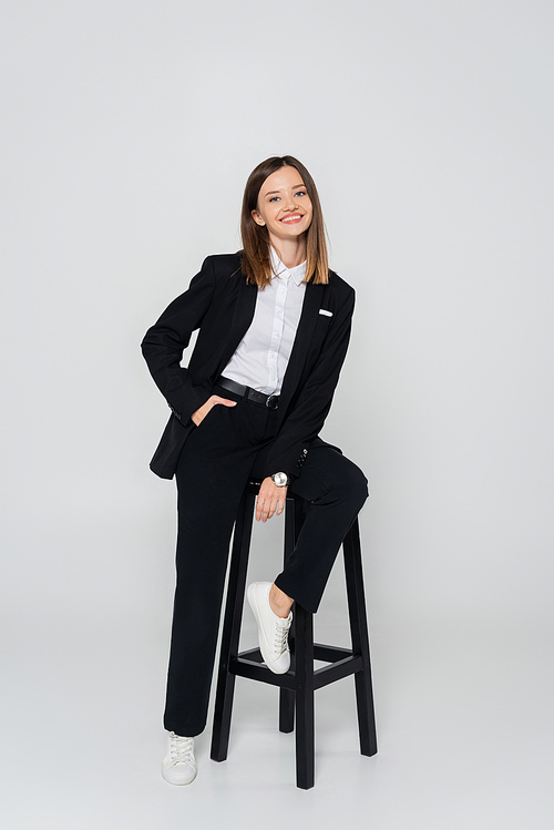 full length of happy woman in suit posing with hand in pocket while sitting chair on grey