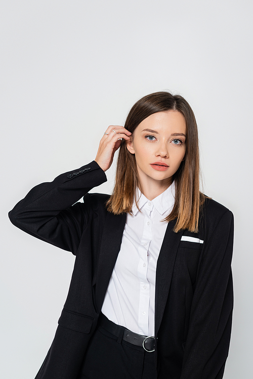 young stylish woman in suit adjusting hair isolated on grey