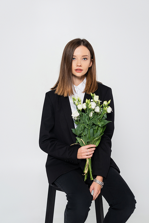 young stylish woman in suit holding bouquet of flowers and sitting on chair isolated on grey