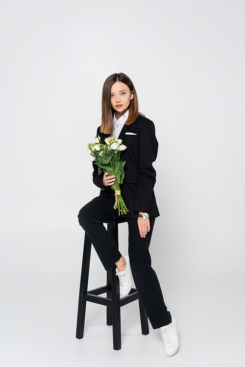 full length of stylish woman in suit holding bouquet of flowers and sitting on chair on grey