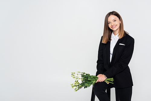 cheerful woman in suit holding bouquet of flowers and sitting on chair isolated on grey