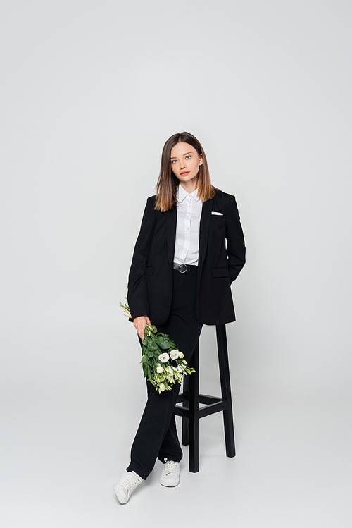 full length of young woman in suit holding bouquet of flowers and leaning on chair on grey