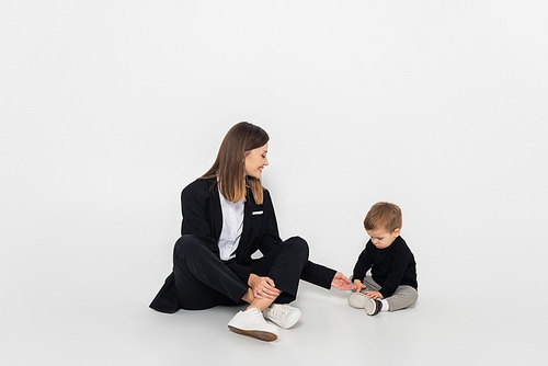 stylish and happy woman sitting near toddler son on grey