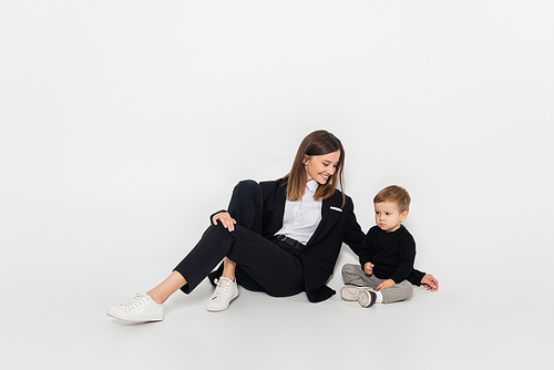 stylish and cheerful woman sitting near toddler son on grey