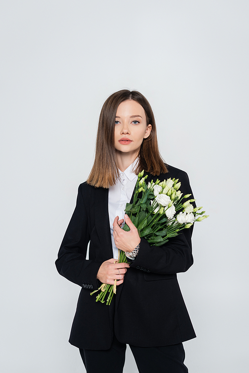 trendy young woman in black suit holding flowers isolated on grey