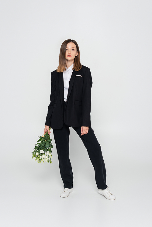 full length of trendy young woman in black suit holding flowers on grey