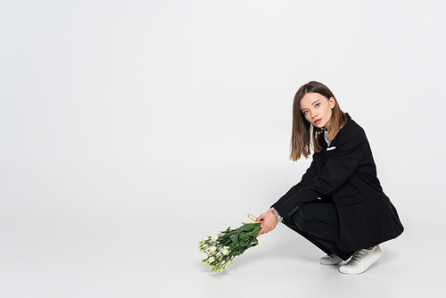 trendy young woman in black suit sitting and holding white flowers on grey
