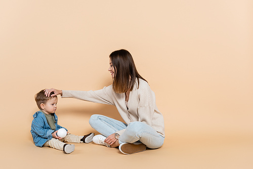 happy mother sitting and adjusting hair of toddler son on beige