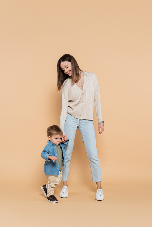 full length of happy mother standing and holding hands with toddler son on beige