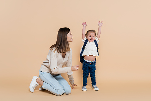 cheerful mother looking at happy child with down syndrome raising hands on beige