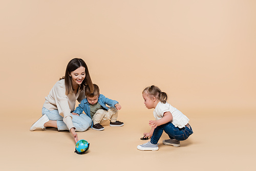 happy woman playing with globe near toddler boy and girl with down syndrome on beige